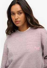 Jasnoróżowa bluza wykonana z miękkiego materiału, z okrągłym dekoltem, długimi rękawami i haftowanym logo na piersi.
