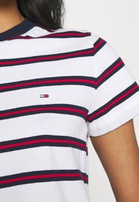 Camiseta blanca de algodón con rayas horizontales en azul marino y rojo, que presenta un pequeño parche con logo en el lado izquierdo del pecho y un cuello de canalé en azul marino.
