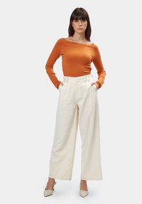 Langärmliges Oberteil aus rostfarbenem Stoff, tailliertem Schnitt. Cremefarbene, weit geschnittene Hose mit Falten und hoher Taille. Weiches Gewebe in neutralen Farbtönen. Beigefarbene Absatzschuhe.