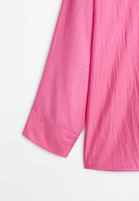 Manche longue d'une chemise rose vif à texture lisse avec un poignet large à un seul bouton sur fond blanc.