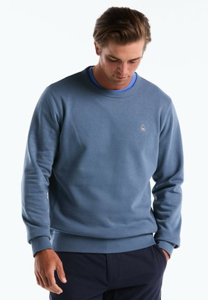 Homme portant un sweat-shirt bleu à col rond avec un petit logo d'animal sur la poitrine, regardant vers le bas avec une main dans sa poche.