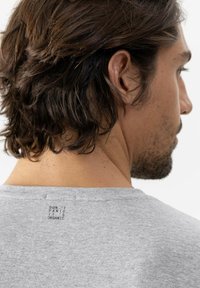 Man met schouderlang, donker haar, draagt een grijs shirt met kleine tekst "DON'T PANIC IT'S ORGANIC" op de bovenrug.