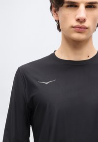 Νεαρός άνδρας που φοράει μαύρο πουκάμισο μακρυμάνικο με ένα μικρό ασημένιο λογότυπο πουλιού στο στήθος, εμφανιζόμενος από τον λαιμό ως το μέσο του κορμού.