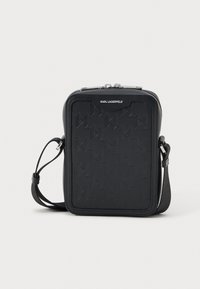 Melnas krustsoma, kas izgatavota no teksturētas ādas, ar ģeometrisku rakstu, ar rāvējslēdzēju un regulējamu siksniņu.