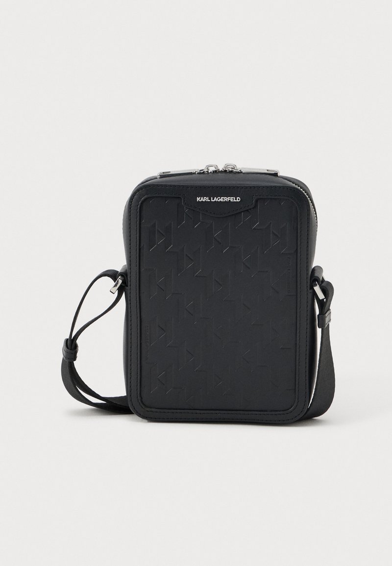 Melnas krustsoma, kas izgatavota no teksturētas ādas, ar ģeometrisku rakstu, ar rāvējslēdzēju un regulējamu siksniņu.