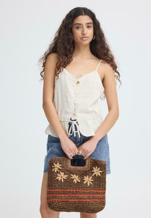 Giovane donna con lunghi capelli ricci, indossa un top crema senza maniche e pantaloncini di jeans, tiene una borsa marrone intrecciata con decorazione floreale.