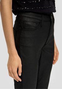 Svarta denimjeans med en åtsittande design, framfickor och en mjuk textur. Nära vy som framhäver midjebandet och handens placering.