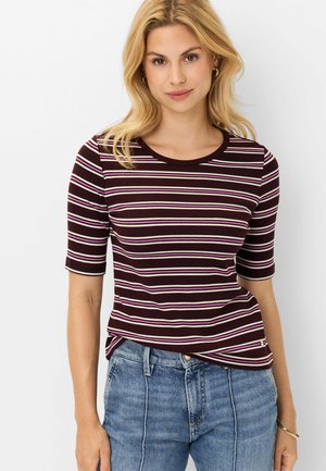 Frau mit blondem Haar trägt ein dunkelviolettes, gestreiftes Kurzarmtop und blaue Jeans, steht vor einem weißen Hintergrund.