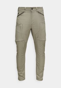 Cargo-byxor i ljus khaki, med flera sidofickor, en figurnära design och kontrasterande mörka kantlinjer längs fickorna.