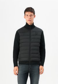 Black puffer vest with a high collar, quilted texture, and contrasting sleeves. Zipper closure and emblem on the chest. Paired with dark jeans.