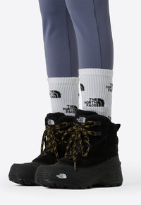 Μαύρα αδιάβροχα μποτάκια με κίτρινες κορδόνια, ανώτερο ύφασμα με υφή και χοντρές σόλες, συνδυασμένα με λευκές κάλτσες που φέρουν μαύρο λογότυπο.