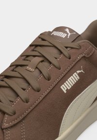 Καφέ σουέντ αθλητικό παπούτσι Puma με καφέ κορδόνια, λευκό λογότυπο Puma στο πλάι και στη γλώσσα, και σόλα και λεπτομέρεια ρίγας σε κρεμ απόχρωση.