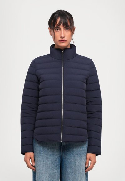 Kvinde iført en lynlåset marineblå quilted jakke og lyseblå jeans mod en ensfarvet hvid baggrund.