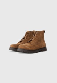 Brune suedes ankelstøvler med flad gummisål, rund tå og snørelukning. Har syninger og en løkke bagpå.