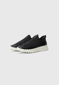 Black slip-on sneakers featuring a textured knit upper, rubberized toe cap, and a chunky white sole with a wavy design.