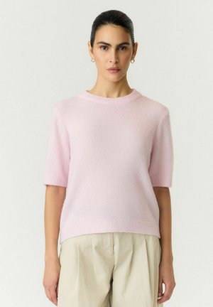 Femme portant un pull rose clair à manches courtes et un pantalon beige, debout et faisant face à l’avant sur un fond clair uni.