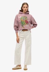 Sudadera con capucha rosa que presenta un gráfico de dragón verde y detalles florales, combinada con pantalones de pierna ancha blancos y zapatillas beige.