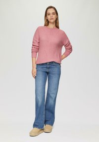 Pull en tricot rose avec un motif texturé, associé à un jean évasé bleu et des chaussures slip-on beiges. Le mannequin se tient devant un fond neutre.