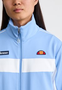 Light blue zip-up jacket with a high collar, white horizontal stripes, and a multicolored embroidered logo on the chest. Smooth fabric texture.