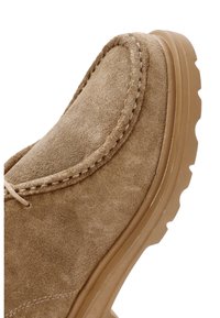 Zapato de ante en tono beige con punta redondeada, costura tonal y suela de goma gruesa y texturizada. El diseño presenta una estética limpia y minimalista.