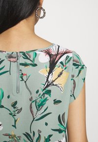 Top con stampa floreale in tessuto verde, con maniche corte, scollatura rotonda e dettaglio con zip sul retro. Orecchini a cerchio in argento visibili.