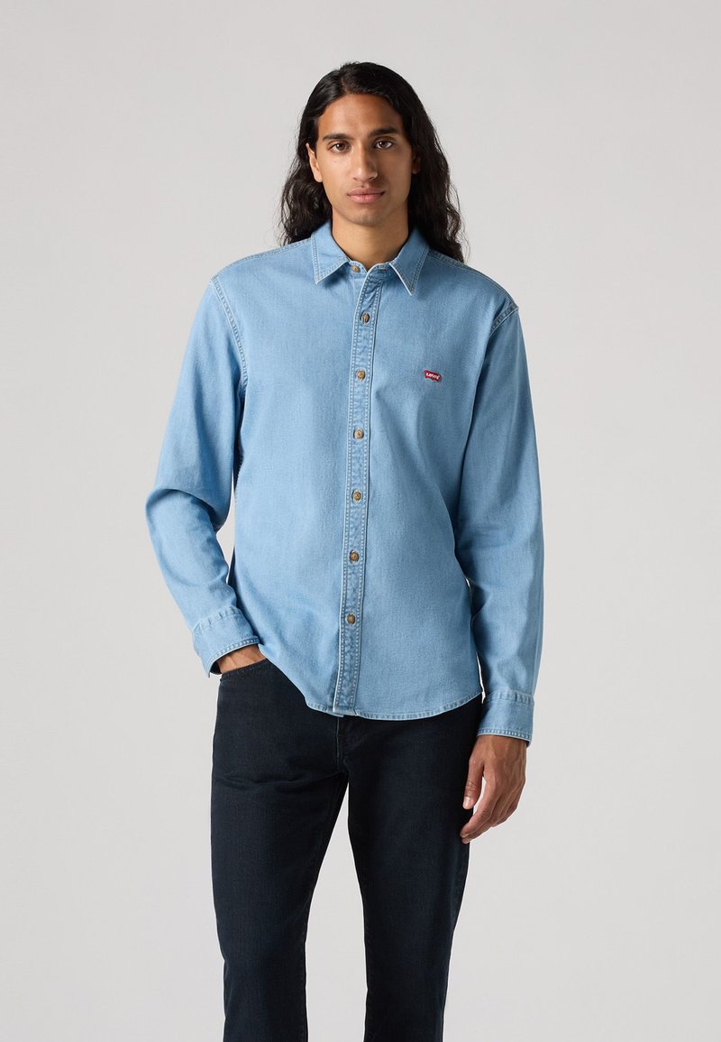 Camicia di jeans azzurro chiaro con colletto, chiusura a bottoni e maniche lunghe. Logo rosso sul lato sinistro del petto. Indossata con jeans scuri.