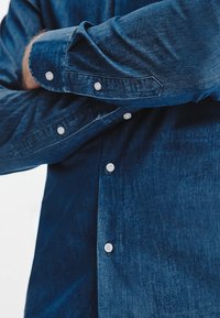 Jeansowa koszula w ciemnoniebieskim kolorze, z zapięciem na guziki i kontrastowymi białymi guzikami. Gładka tekstura z lekko wyblakłym wyglądem.