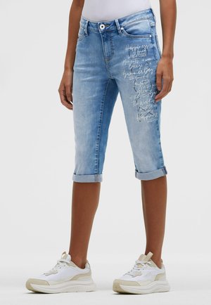 Person trägt hellblaue, knielange Jeansshorts mit umgekrempeltem Saum und weißem Schriftzug auf dem linken Oberschenkel, weiße Sneaker und ein weißes Oberteil.