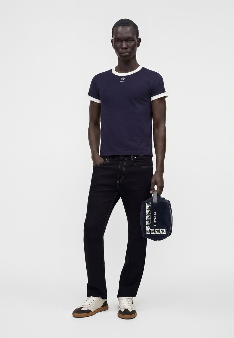 Homme portant un T-shirt bleu marine avec des bordures blanches, un jean noir, des baskets blanches et marron, tenant un sac Versace bleu marine avec un motif blanc.