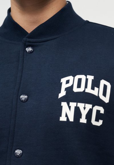 Κοντινό πλάνο σκούρας μπλε μπουφάν με κουμπιά τύπου τρουκ και λευκό κείμενο που γράφει "POLO NYC" στο στήθος.