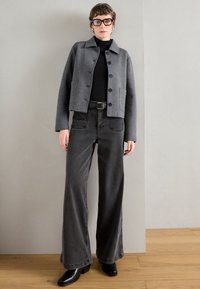 Casaco cinza cortado com botões, suéter preto de gola alta e jeans pretos de perna larga de cintura alta. Botas pretas de salto bloco completam o visual.