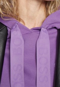 Sudadera morada con un tejido texturizado, un bolsillo frontal y cordones de logotipo a tono. El diseño incluye un acento de cuero negro.