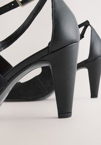 Czarne sandały na wysokim obcasie wykonane ze skóry o gładkiej fakturze, z regulowanym paskiem na kostkę i rzeźbionym, blokowym obcasem z wycięciami.