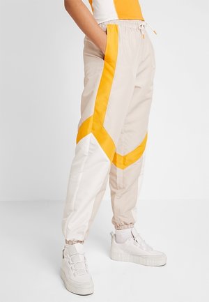 Pantalon jogger beige clair avec des rayures géométriques jaunes et blanches, poignets élastiques, associé à des baskets blanches et des chaussettes blanches.