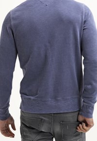 Modrý, texturovaný svetr s výstřihem do V a dlouhými rukávy. Švy na ramenech a v dolním lemu. Nosený přes šedé džíny, ruce v kapsách.