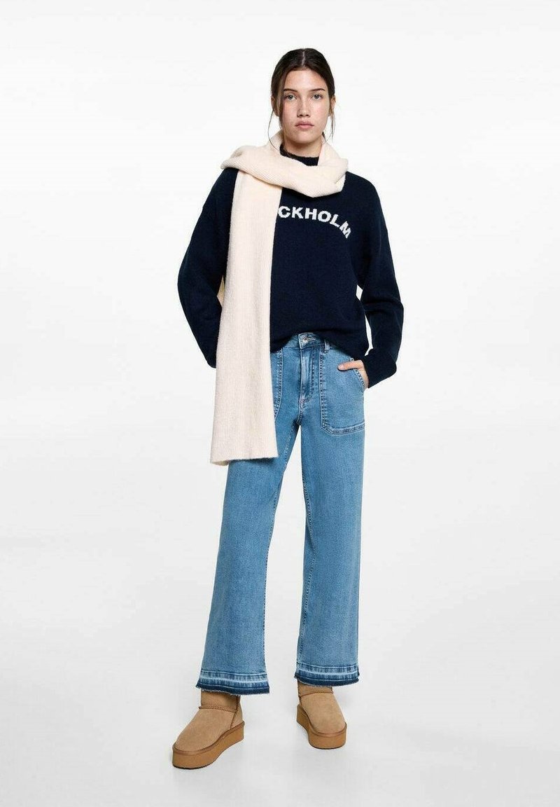 Marinblå tröja med "STOCKHOLM" grafik, krämfärgad scarf, ljusblå vida jeans och beige plattformstövlar. Texturen är mjuk, materialen varierar.