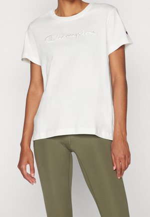 Femme portant un t-shirt blanc Champion avec logo embossé et des leggings vert olive, montrée du cou à la mi-cuisse, sur fond uni.