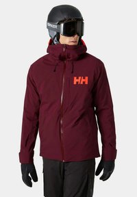 Skijack in bordeaux stof met een hoge kraag, voorrits en rood logo. Voorzien van verstelbare manchetten en zwarte handschoenen. Matte afwerking.