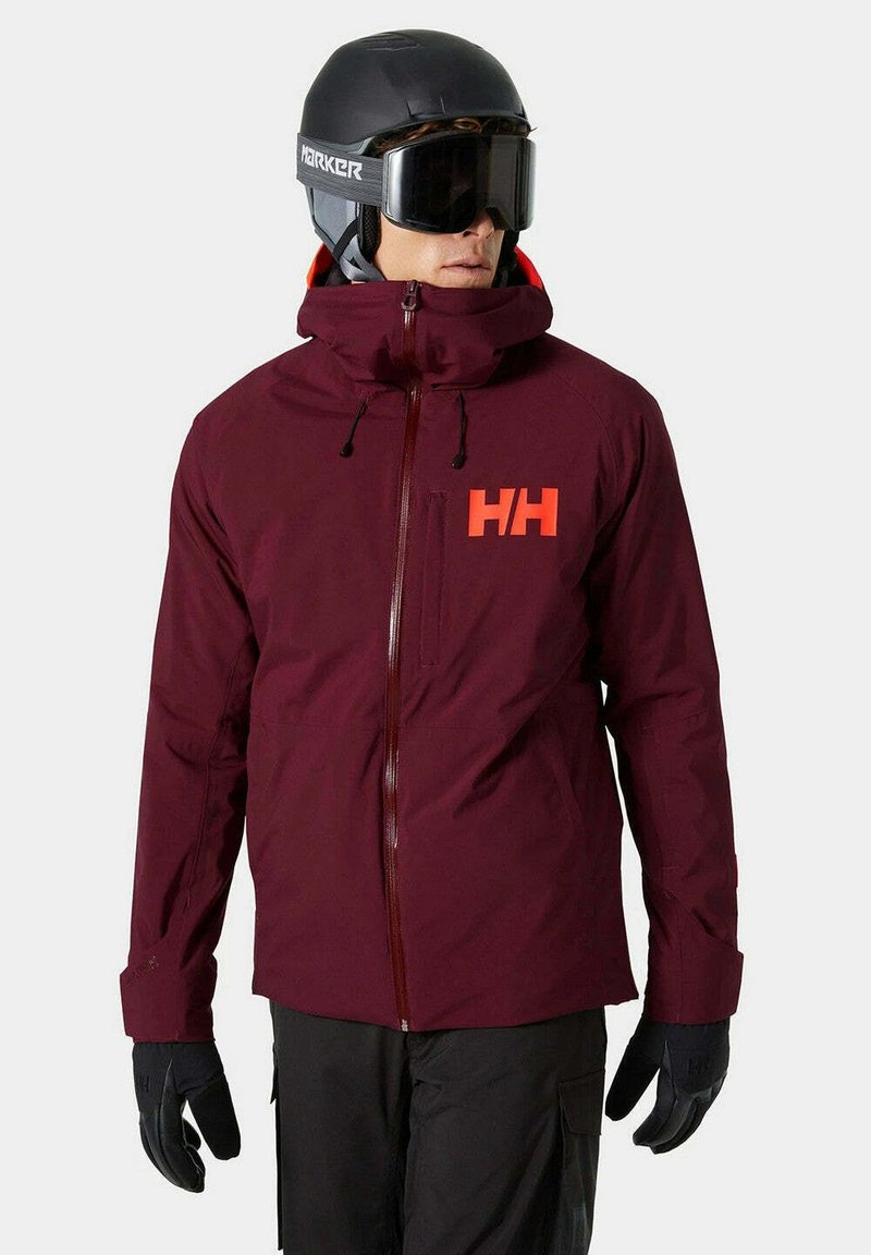 Skijack in bordeaux stof met een hoge kraag, voorrits en rood logo. Voorzien van verstelbare manchetten en zwarte handschoenen. Matte afwerking.
