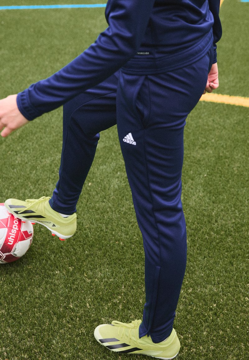Námornícke športové nohavice so bočnými pruhmi, v kombinácii so žiarivo žltými futbalovými topánkami. Hráč sa zaoberá červeným futbalovým loptou na umelom trávniku.
