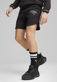 Pantalones cortos deportivos negros con el logo de PUMA, combinados con zapatillas negras y calcetas negras que tienen rayas blancas. Tela suave, corte relajado.