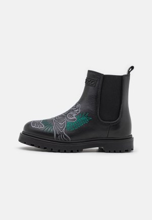 Botte Chelsea en cuir noir avec panneaux latéraux élastiques, arborant un motif de tigre brodé en vert et gris, et une semelle en caoutchouc texturée.