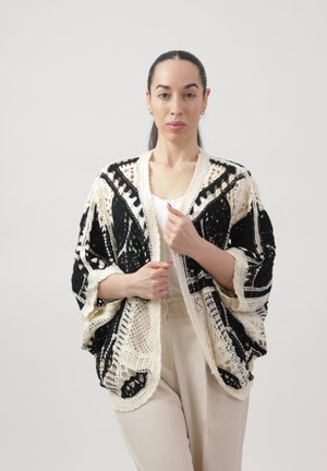 Donna con capelli scuri raccolti in una coda di cavallo che indossa un cardigan in crochet bianco e nero sopra una maglietta bianca e pantaloni beige, in piedi davanti a uno sfondo uniforme.