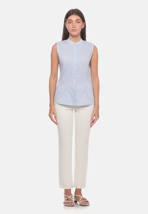 Donna in piedi con una camicetta senza maniche a righe azzurro chiaro, pantaloni bianchi e sandali bianchi con cinturini su uno sfondo semplice.