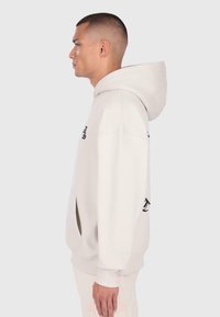 Mladý muž s krátko zostrihnutými vlasmi, oblečený v svetlo béžovej mikine s kapucňou, stojí v profilu na jednofarebnom pozadí.