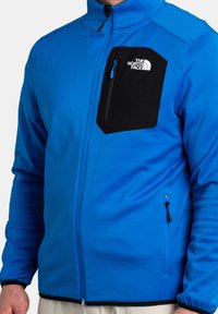 Veste en polaire bleue avec une poche poitrine noire à zip et logo, poches latérales zippées et poignets ajustés. Texture douce et design flexible.