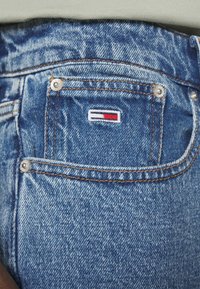 Džíny z denimu s modrým přelivem, měděným prošitím a malým vyšitým logem s červenými, bílými a modrými akcenty na zadní kapse.