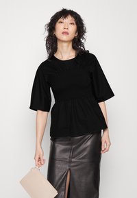 Top peplum preto com corpete franzido e mangas largas, combinado com uma saia de couro preta brilhante com uma fenda frontal, e segurando uma carteira bege.