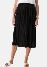 Jupe plissée noire en tissu léger, dotée d'une taille élastique et d'une longueur mi-mollet. Portée avec des sandales blanches à enfiler.