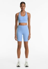 Svijetloplavi atletski crop top s širokim remenima u kombinaciji s odgovarajućim visokim šortsevima, izrađen od glatkog, rastezljivog materijala.